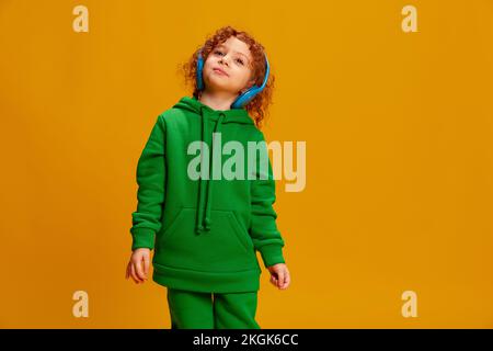 Portrait of cute little girl, child with curly red hair posing in headphones isolated over yellow background Stock Photo