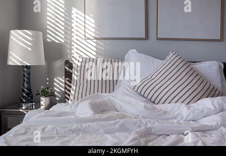 Early morning light casts light and shadows from window blinds onto striped accent pillows and blue and white covers on a bed Stock Photo