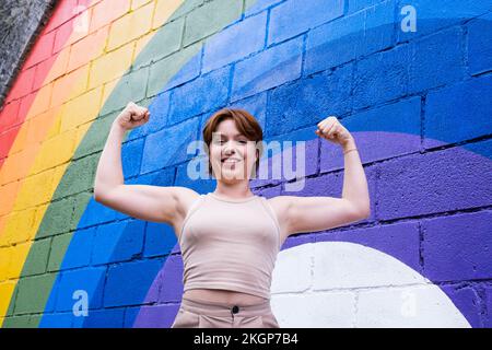Happy young woman flexing muscles in front of rainbow flag painted on wall Stock Photo