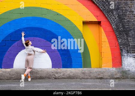 Young woman with hand raised leaning on rainbow flag painted on wall Stock Photo
