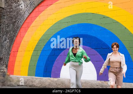 Happy woman with friend running in front of rainbow colored wall Stock Photo