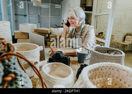 Smiling craftswoman talking on mobile phone and using laptop on workbench Stock Photo