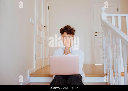 Boy with hands clasped sitting with laptop on stairs at home Stock Photo