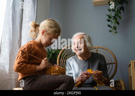 Smiling senior woman looking at granddaughter knitting in living room Stock Photo