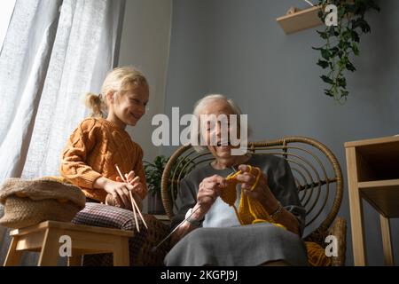 Happy girl holding ball of wool and needle looking at grandmother knitting on chair Stock Photo