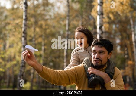 Playful man playing with paper airplane carrying son on shoulders in park Stock Photo