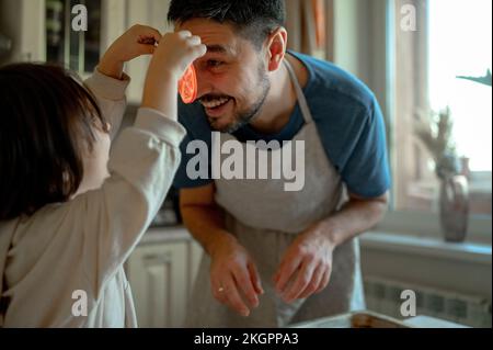 Happy father and son playing with tomato slices in kitchen Stock Photo