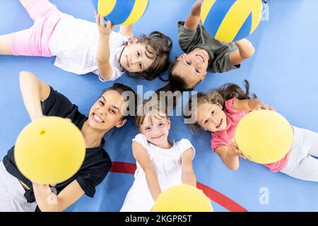 Happy students and teacher lying down with ball at school sports court Stock Photo