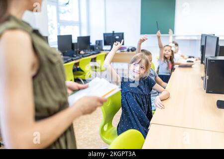 Students with hand raised looking at teacher standing in class at school Stock Photo