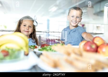 Smiling elementary students taking fruits at lunch break in school cafeteria Stock Photo