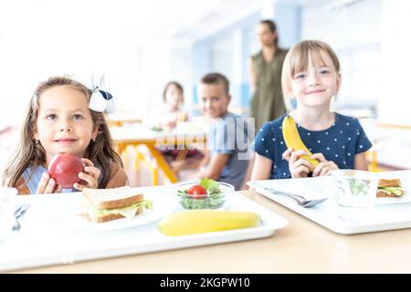 Girls holding apple and banana sitting at table in cafeteria Stock Photo