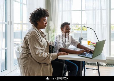 Mother sitting by son doing homework using laptop on table at home Stock Photo