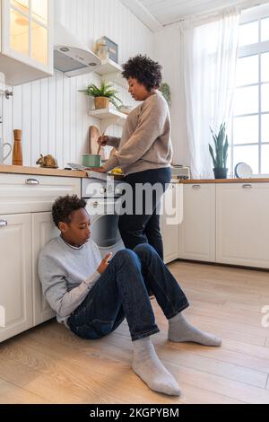 Mother preparing food with son sitting on floor by cabinet in kitchen at home Stock Photo