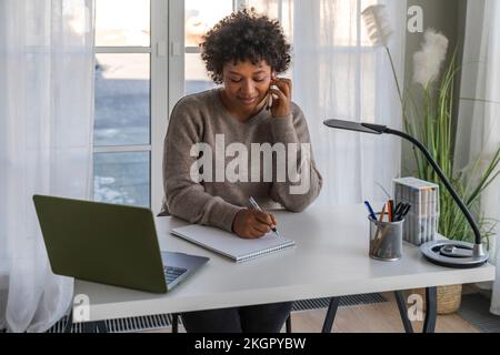 Freelancer talking through smart phone and taking down notes at home Stock Photo