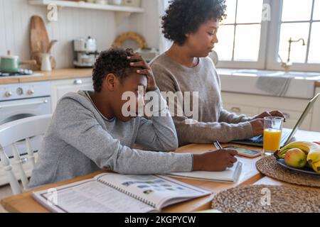 Stressed boy doing homework by mother using laptop at table Stock Photo