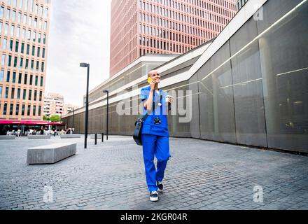 Smiling nurse talking over mobile phone and walking on footpath Stock Photo