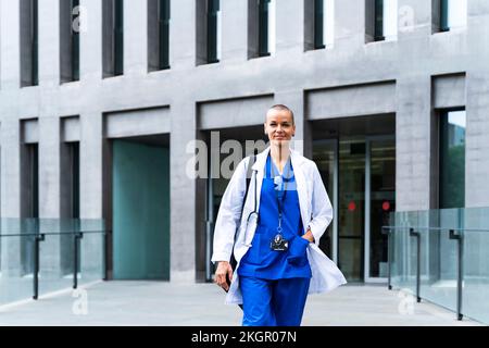Smiling female doctor with hand in pocket walking outside hospital Stock Photo