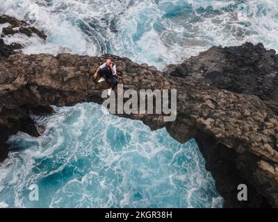 Mature man sitting on rock arch above sea Stock Photo