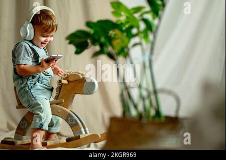 Happy boy wearing wireless headphones using smart phone on rocking horse Stock Photo