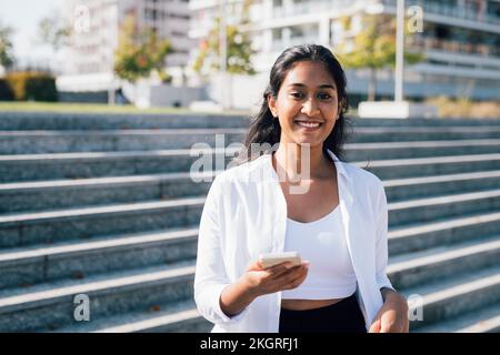 Smiling young woman with smart phone in front of steps Stock Photo