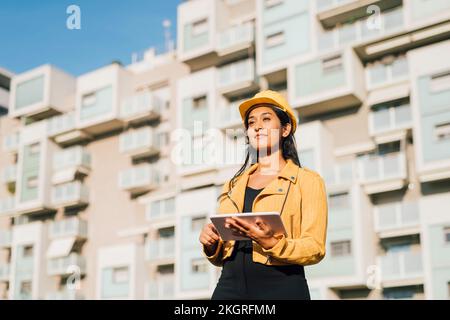 Contemplative young woman wearing hardhat standing in front of building Stock Photo