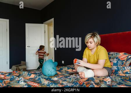 Boy with plaster cast on leg playing with puzzle cube sitting on bed at home Stock Photo