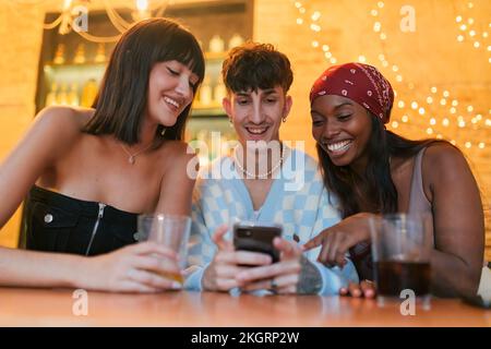 Happy young man sharing smart phone sitting amidst friends in restaurant Stock Photo