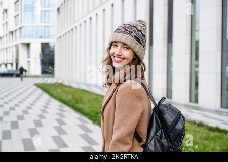 Happy woman wearing knit hat standing with backpack on footpath Stock Photo