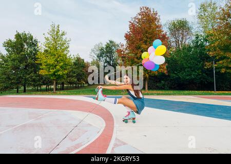 Happy young woman holding colorful balloons roller skating at sports court Stock Photo