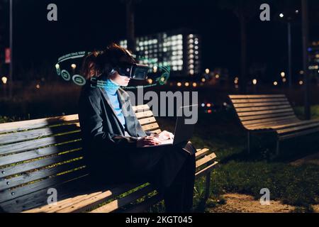 Woman wearing VR glasses using laptop on bench at night Stock Photo
