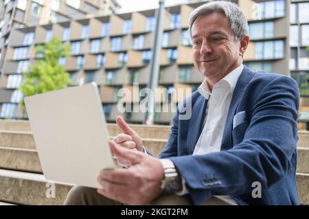 Smiling mature man showing thumbs up on video call using laptop at stairs Stock Photo