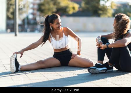 Teenage girl exercising and talking to friend sitting on footpath Stock Photo