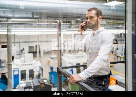 Young businessman talking on smart phone holding railing at factory Stock Photo