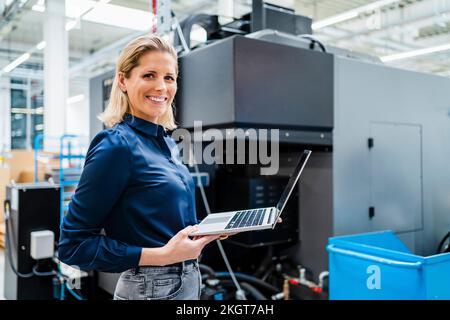 Smiling businesswoman with laptop standing by machinery in factory Stock Photo