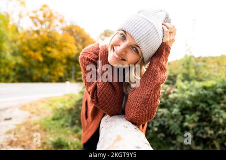 Smiling woman wearing knit hat leaning on wood in autumn Stock Photo
