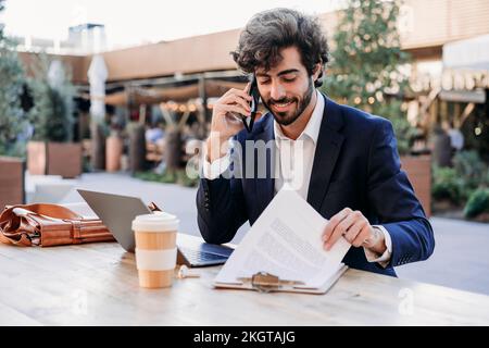 Businessman reviewing documents and talking on smart phone at table Stock Photo