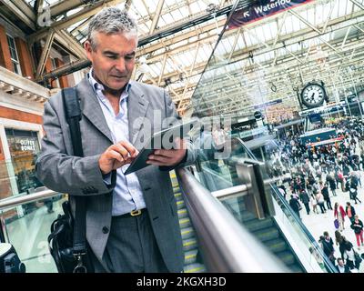 iPad Businessman using smart tablet on busy commuter commuting rail Waterloo Station railway concourse with clock, business on the move.  London UK Stock Photo