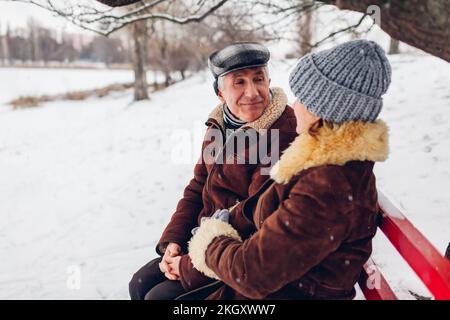 Senior family couple sitting on bench outdoors during snowy winter weather. Elderly retired people chatting in park. Valentine's day Stock Photo