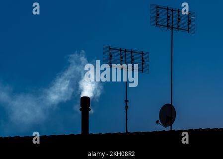 Chimney with white smoke, tv antennas and satellite dish seen in silhouette on rooftop, copy space Stock Photo