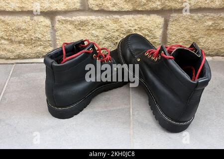 Black walking boots with red laces awaiting cleaning on grey tiled floor with stone coloured bricks to the rear, after a day walking in the outdoors. Stock Photo