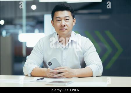 Portrait of a serious young Asian freelancer, designer, architect. He sits at the desk in the office, holds the phone, works with documents, looks into the camera. Stock Photo