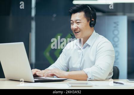 Learning online. Young handsome male Asian student studying remotely with laptop and headphones. Sitting at a desk in an office on campus. Stock Photo