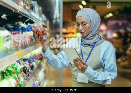 A Muslim woman in a hijab, a supermarket worker checks products on shelves and in refrigerators, a woman with a tablet and an apron uses a tablet to review products. Stock Photo