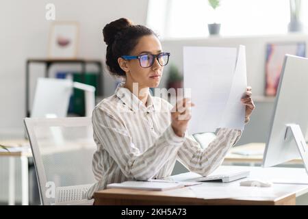 Serious busy woman in glasses sitting at table with computer and working with documents in office interior Stock Photo