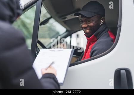 Happy positive Black courier joking around with unrecognizable person taking a delivery. Unrecognizable person signing delivery documents standing next to white delivery truck. High quality photo Stock Photo