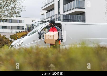 Postal delivery process. Silhouette of tall, well-build Black courier man in red jacket standing with a cardboard parcel in front of white van. Blurred bushes in the foreground. High quality photo Stock Photo