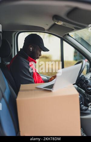 Vertical in-car shot of focused Black middle-aged man in black hat and red-and-gray jacket sitting behind the wheel of package delivery truck. Blurred silver laptop standing on a cardboard in the foreground. High quality photo Stock Photo