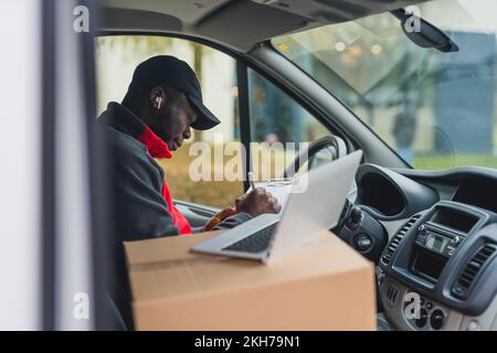 Online shopping delivery process. Focused hard-working African-American man in his 30s sitting on driver's seat behind the wheel of a big white delivery van, writing on his clipboard. Silver laptop and cardboard packaging in the foreground. High quality photo Stock Photo