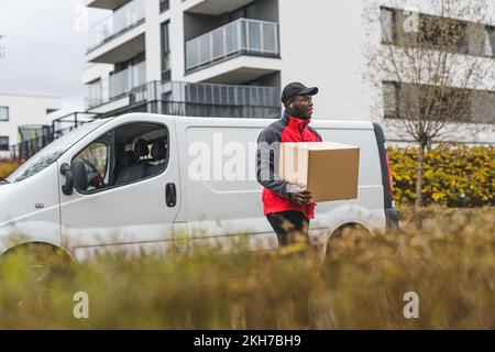 The use of vehicles in package delivery. Outdoor shot of handsome Black deliveryman in front of his white truck walking towards client's house. Blurred foreground. High quality photo Stock Photo