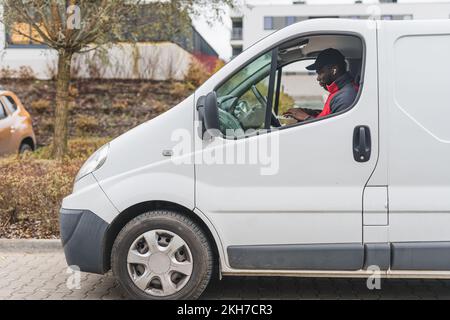 Job - postal delivery person. Outdoor view of a half of white delivery truck and focused Black male courier sitting behind the wheel and working on his laptop. High quality photo Stock Photo